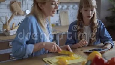 妈妈和女儿一起在一个明亮的大厨房做饭。 妈妈切多汁的辣椒，一个女孩在平板电脑上读到一个食谱